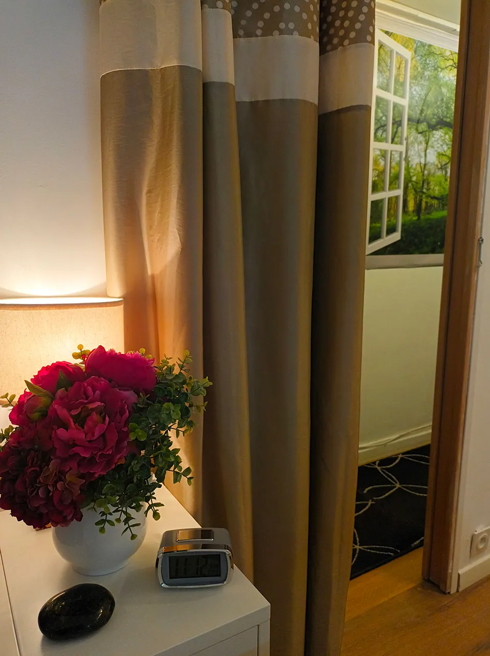 Un coin du cabinet : bouquet de pivoines et lampe près d’un rideau, avec en arrière-plan le poster d’une fenêtre ouverte sur une forêt